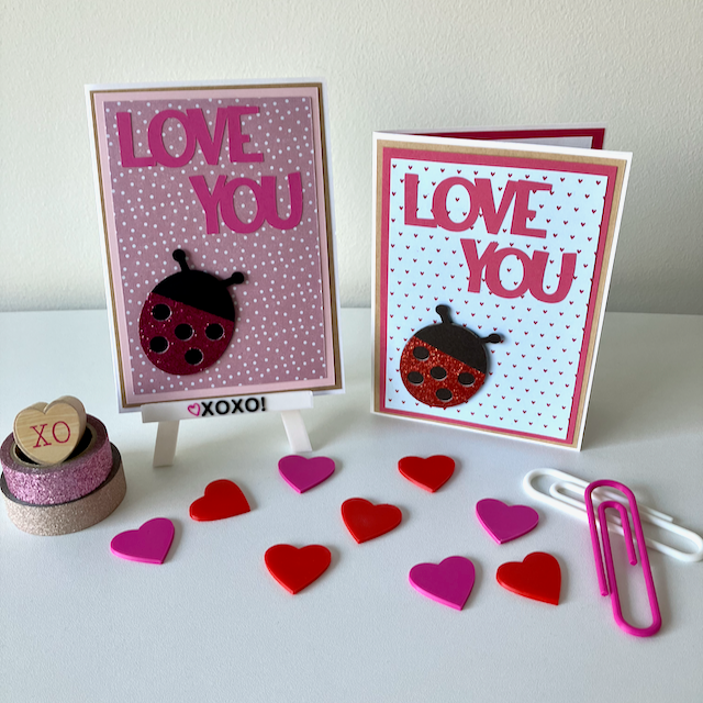 Two handmade greeting cards with lady bugs and 'love you' text displayed on a table with decorative hearts.