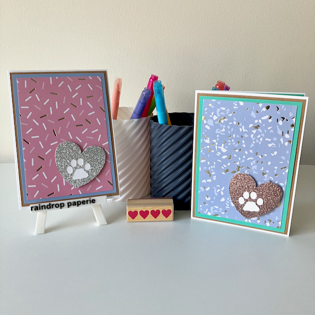 Two handmade greeting cards with paw print designs on a white surface with stationery items.