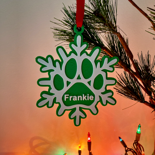 A 3D printed green paw print snowflake ornament with 'Frankie' text hanging on a Christmas tree branch with lights.
