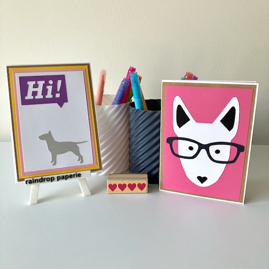 Two handmade greeting cards with English Bull Terriers on a white surface with stationery items. 