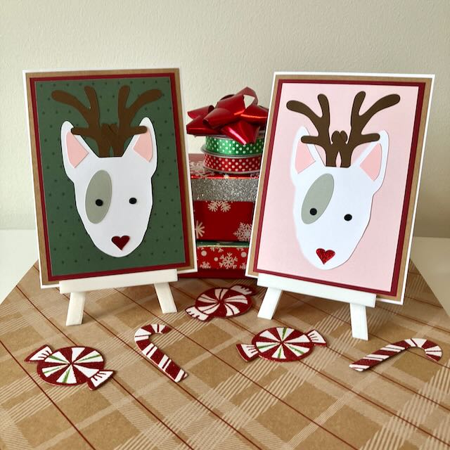 Two handmade Christmas-themed greeting cards with English bull terriers as reindeers on little stands, surrounded by candy canes and gift boxes.