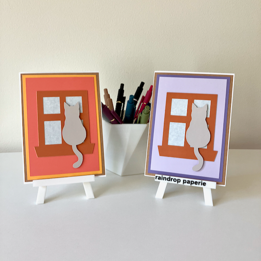 Two handmade greeting cards on stands with cats sitting in window sills. 