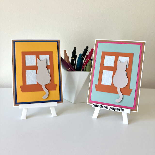 Two handmade greeting cards on stands with cats sitting in window sills. 