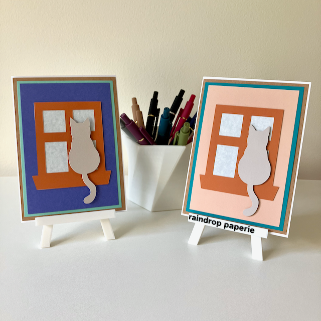 Two handmade greeting cards on stands with cats sitting in window sills. 