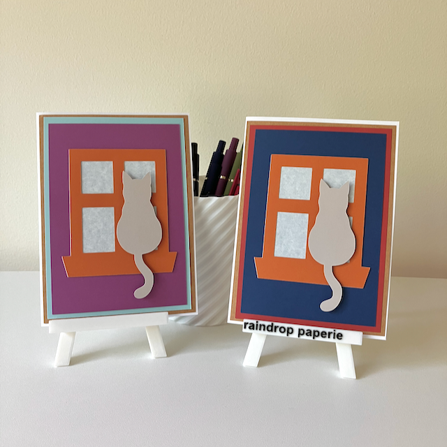 Two handmade greeting cards on stands with cats sitting in window sills. 