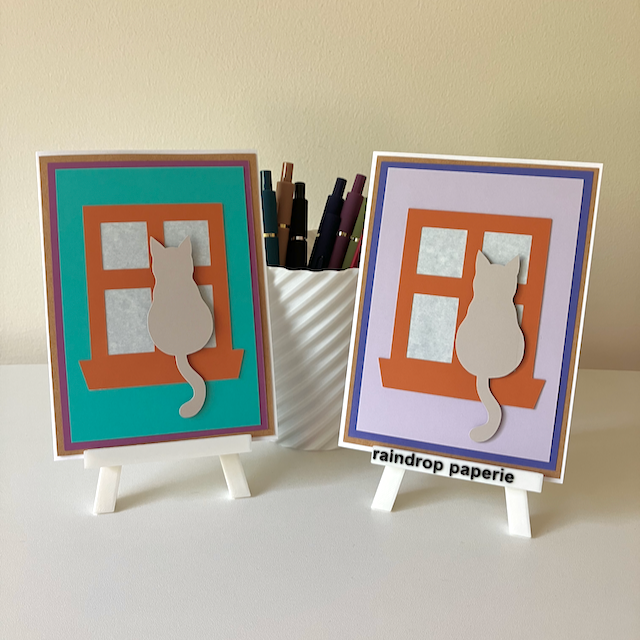 Two handmade greeting cards on stands with cats sitting in window sills. 