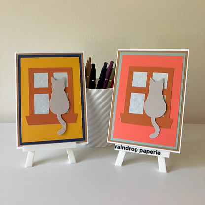 Two handmade greeting cards on stands with cats sitting in window sills. 