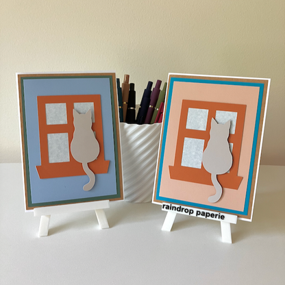 Two handmade greeting cards on stands with cats sitting in window sills. 