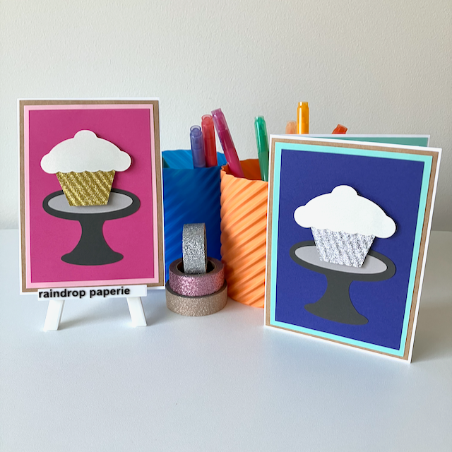 Two greeting cards with cupcake design on stands, surrounded by colorful markers and tape.