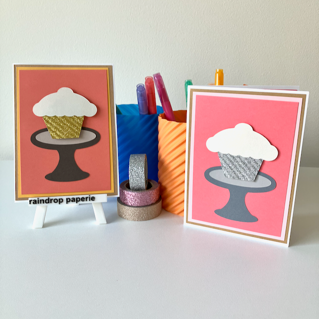 Two greeting cards with cupcake design on stands, surrounded by colorful markers and tape.