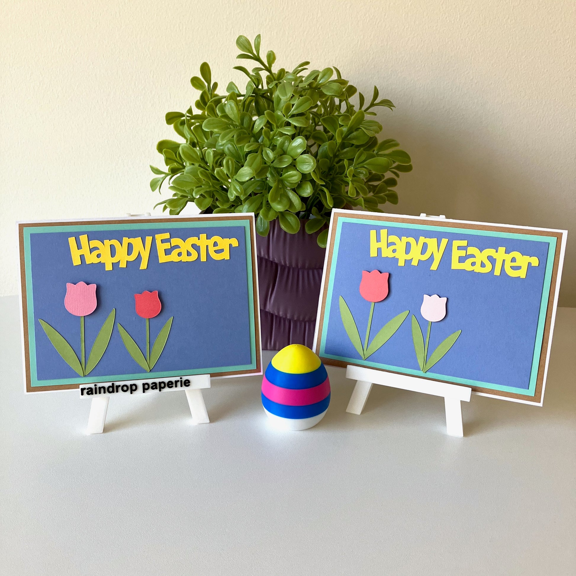 Two Happy Easter greeting cards with tulips displayed on easels with a plant in the background.