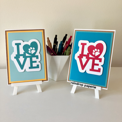 Two handmade greeting cards on stands with a cup of pens. The cards say "LOVE".