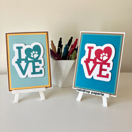 Two handmade greeting cards on stands with a cup of pens. The cards say "LOVE".