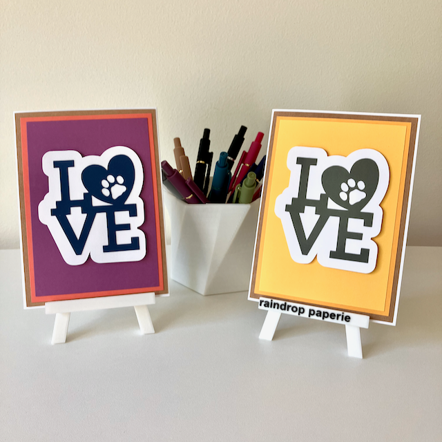 Two handmade greeting cards on stands with a cup of pens. The cards say "LOVE".
