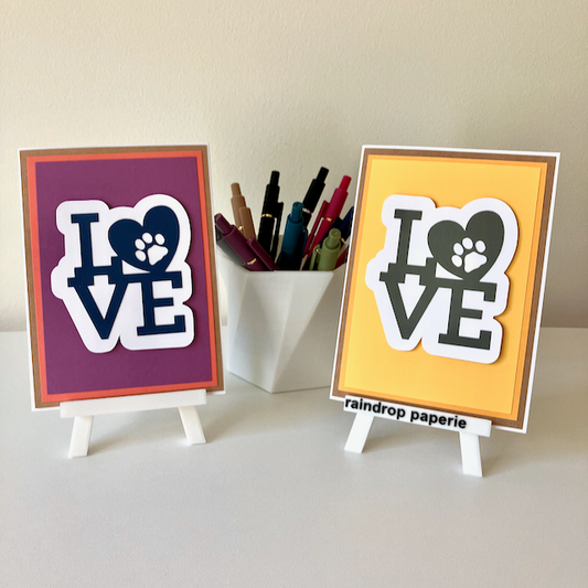 Two handmade greeting cards on stands with a cup of pens. The cards say "LOVE".
