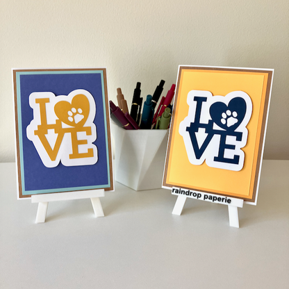 Two handmade greeting cards on stands with a cup of pens. The cards say "LOVE".
