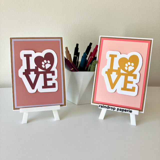 Two handmade greeting cards on stands with a cup of pens. The cards say "LOVE".