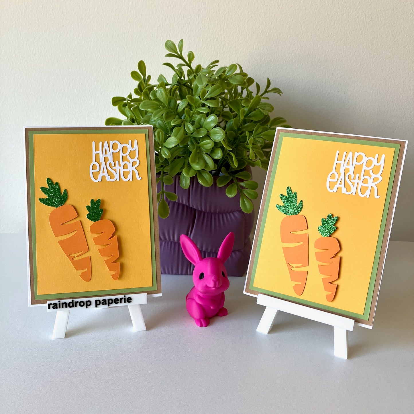 Two Happy Easter greeting cards with carrots displayed on easels with a plant in the background.