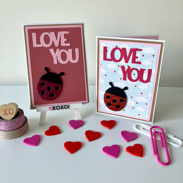 Two handmade greeting cards with lady bugs and 'love you' text displayed on a table with decorative hearts. 