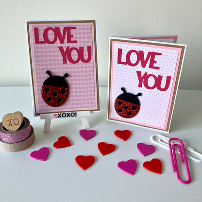 Two handmade greeting cards with lady bugs and 'love you' text displayed on a table with decorative hearts. 