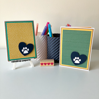 Two handmade greeting cards with paw print designs on a white surface with stationery items.