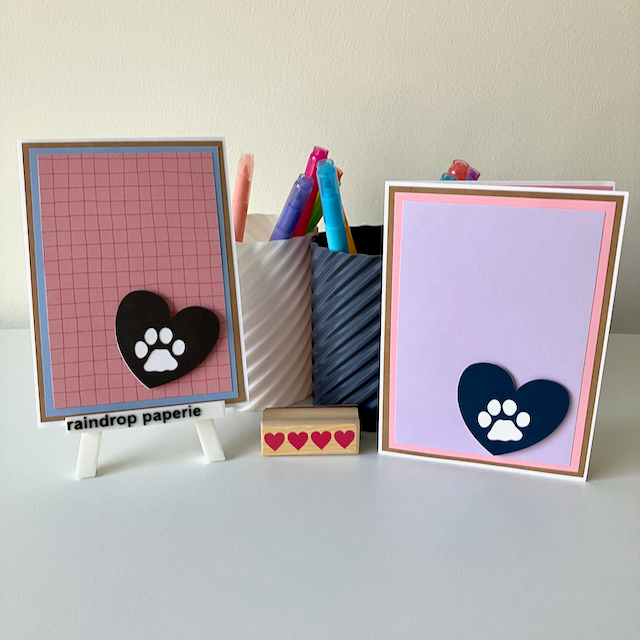 Two handmade greeting cards with paw print designs on a white surface with stationery items.