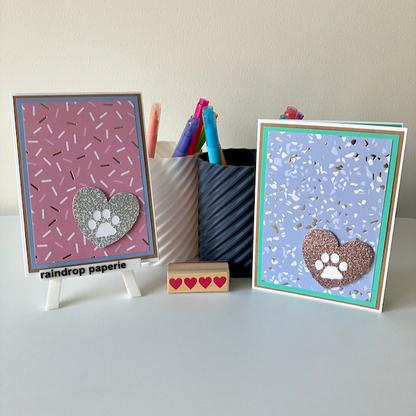 Two handmade greeting cards with paw print designs on a white surface with stationery items.
