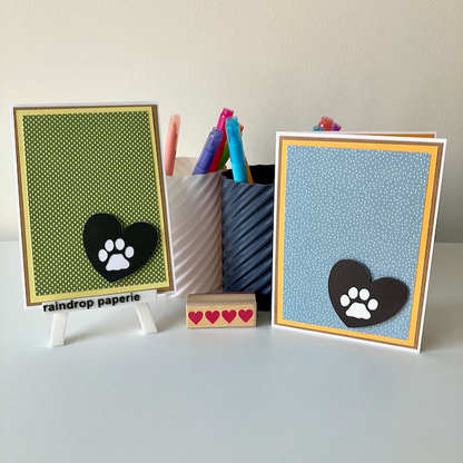 Two handmade greeting cards with paw print designs on a white surface with stationery items.