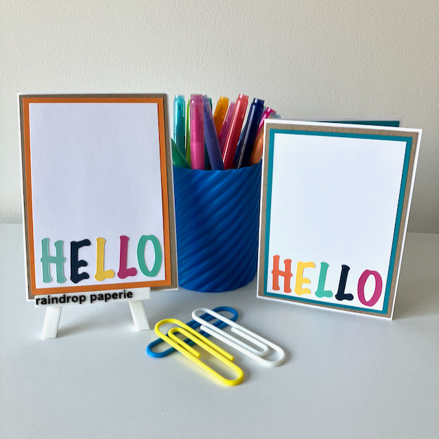 Two handmade greeting cards with 'Hello' text in different colors, placed on a table with a blue pot of pens and paper clips. 