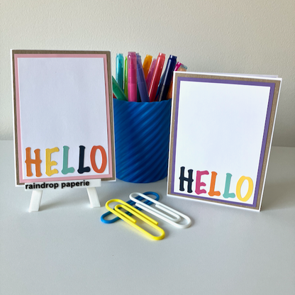 Two handmade greeting cards with 'Hello' text in different colors, placed on a table with a blue pot of pens and paper clips. 