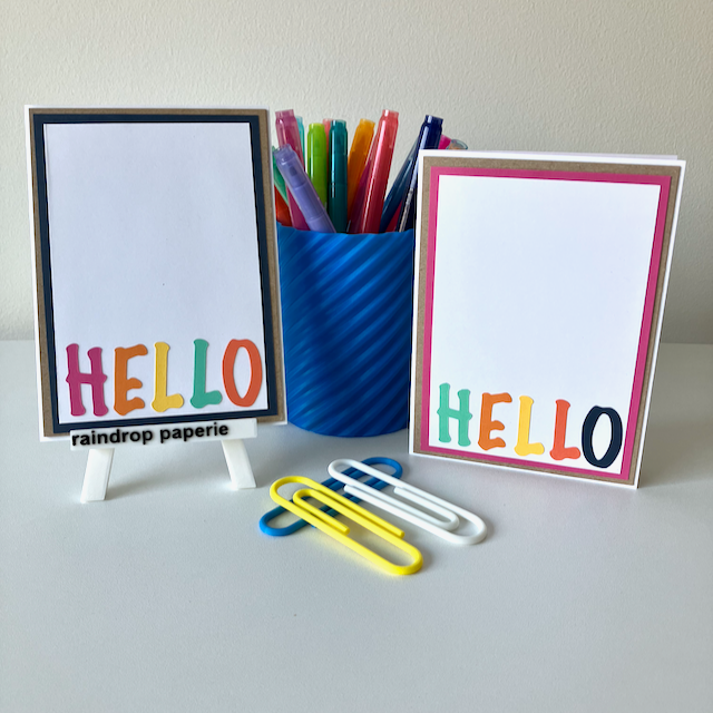 Two handmade greeting cards with 'Hello' text in different colors, placed on a table with a blue pot of pens and paper clips. 