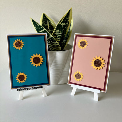 Two sunflower-themed cards on small stands with a plant in the background.
