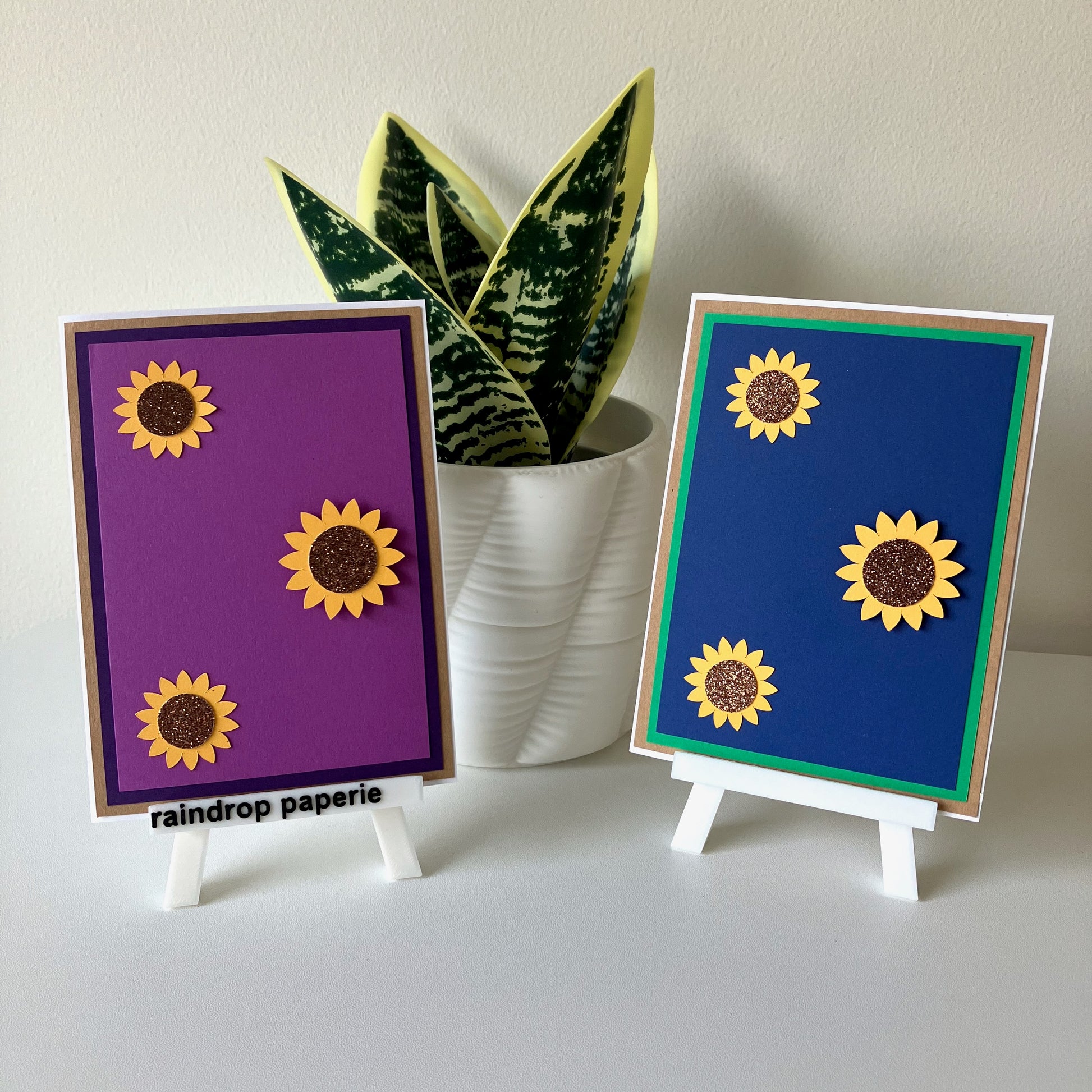 Two sunflower-themed cards on small stands with a plant in the background.