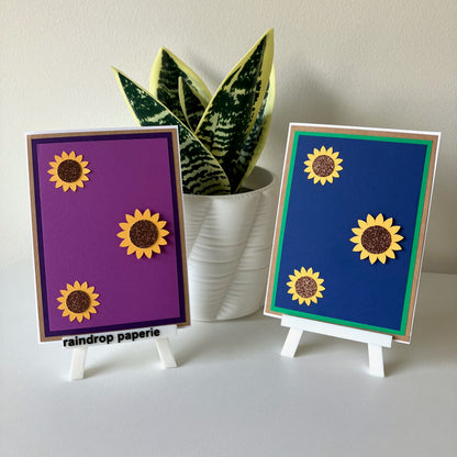 Two sunflower-themed cards on small stands with a plant in the background.
