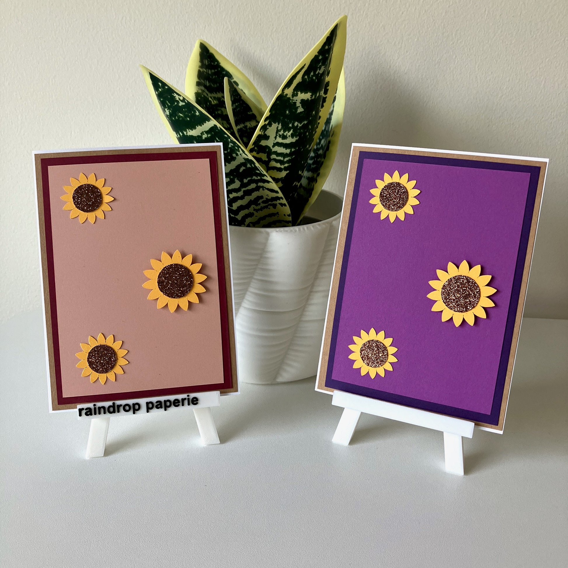Two sunflower-themed cards on small stands with a plant in the background.