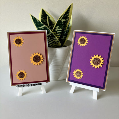Two sunflower-themed cards on small stands with a plant in the background.