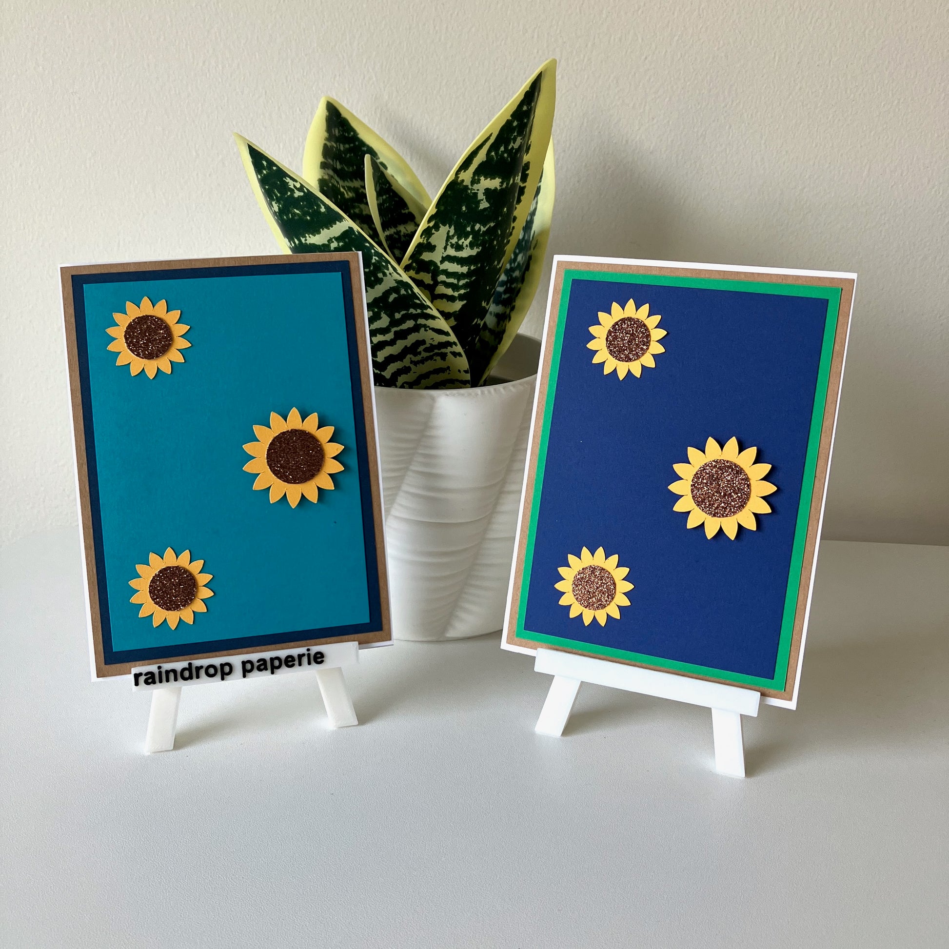 Two sunflower-themed cards on small stands with a plant in the background.